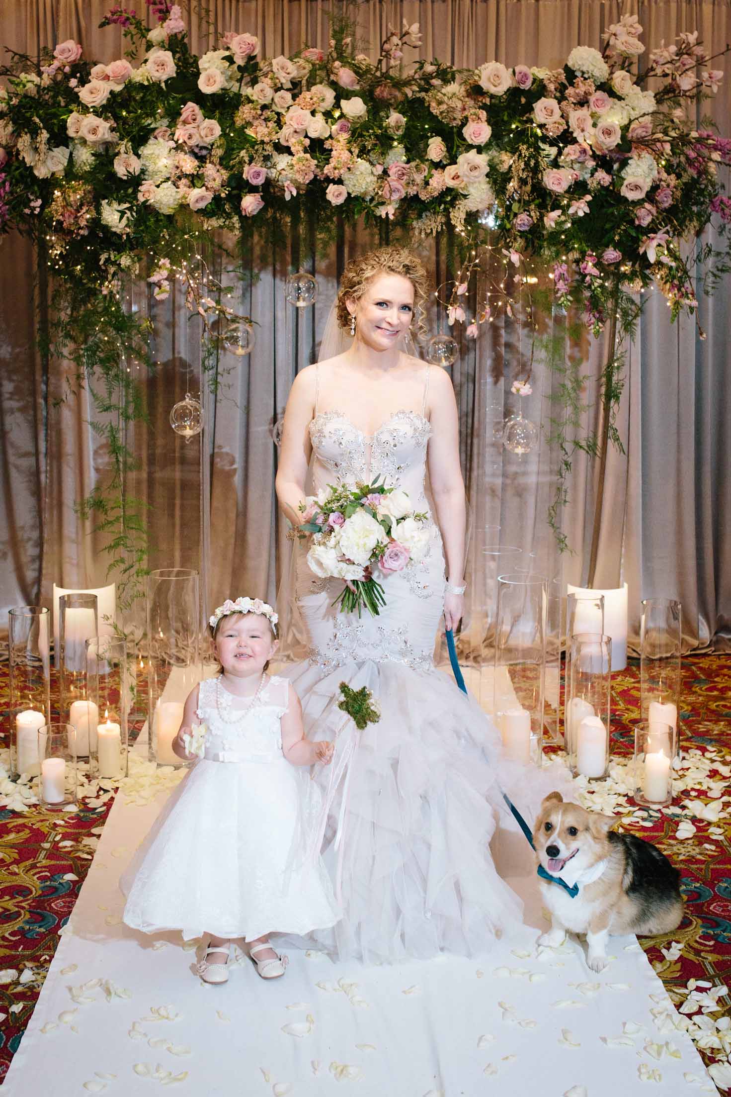 Bride standing with flower girl and dog in front of lush floral spring garden ceremony arch surrounded by candles