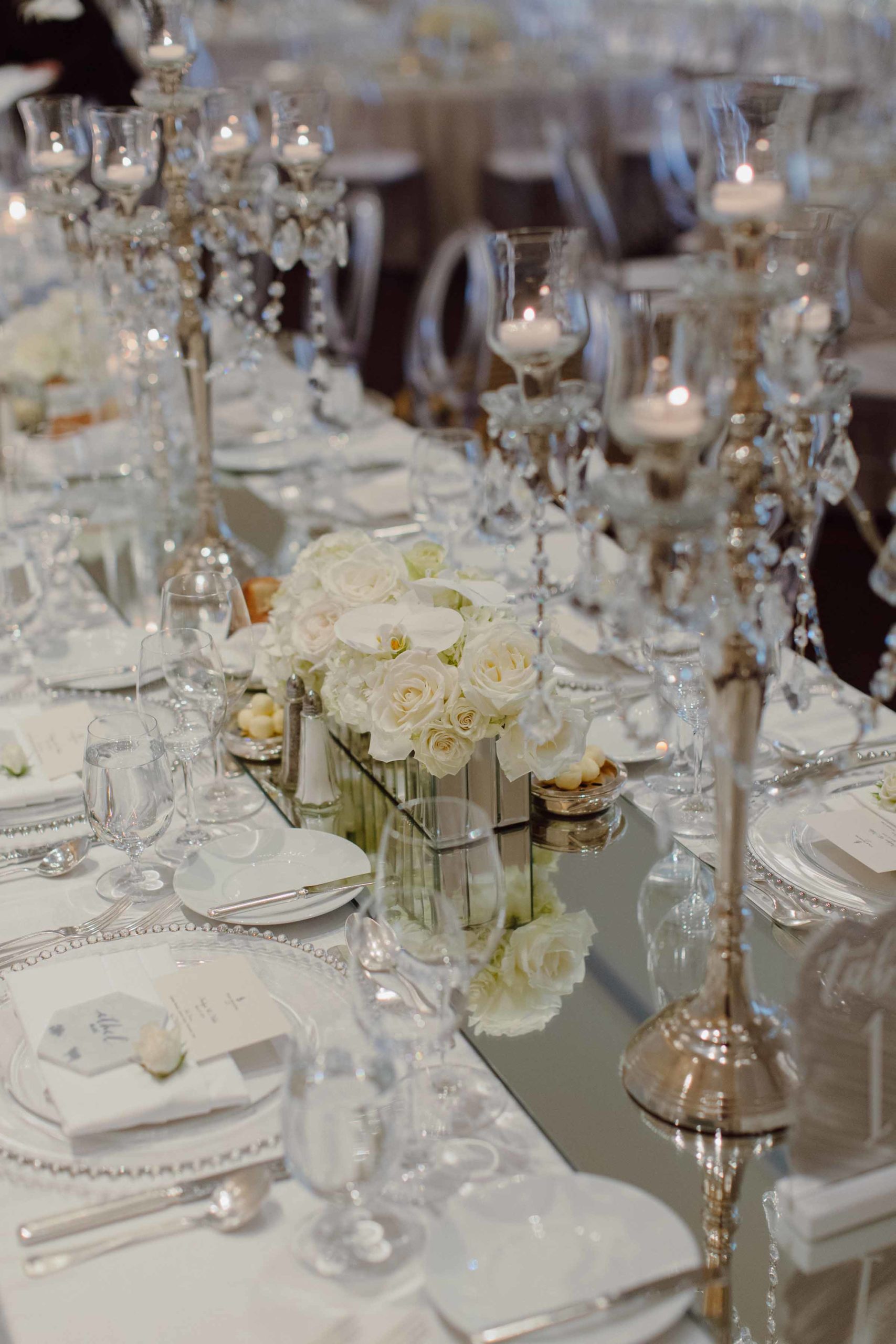 Long wedding reception table with mirrored runner, crystal candelabras, and all white roses