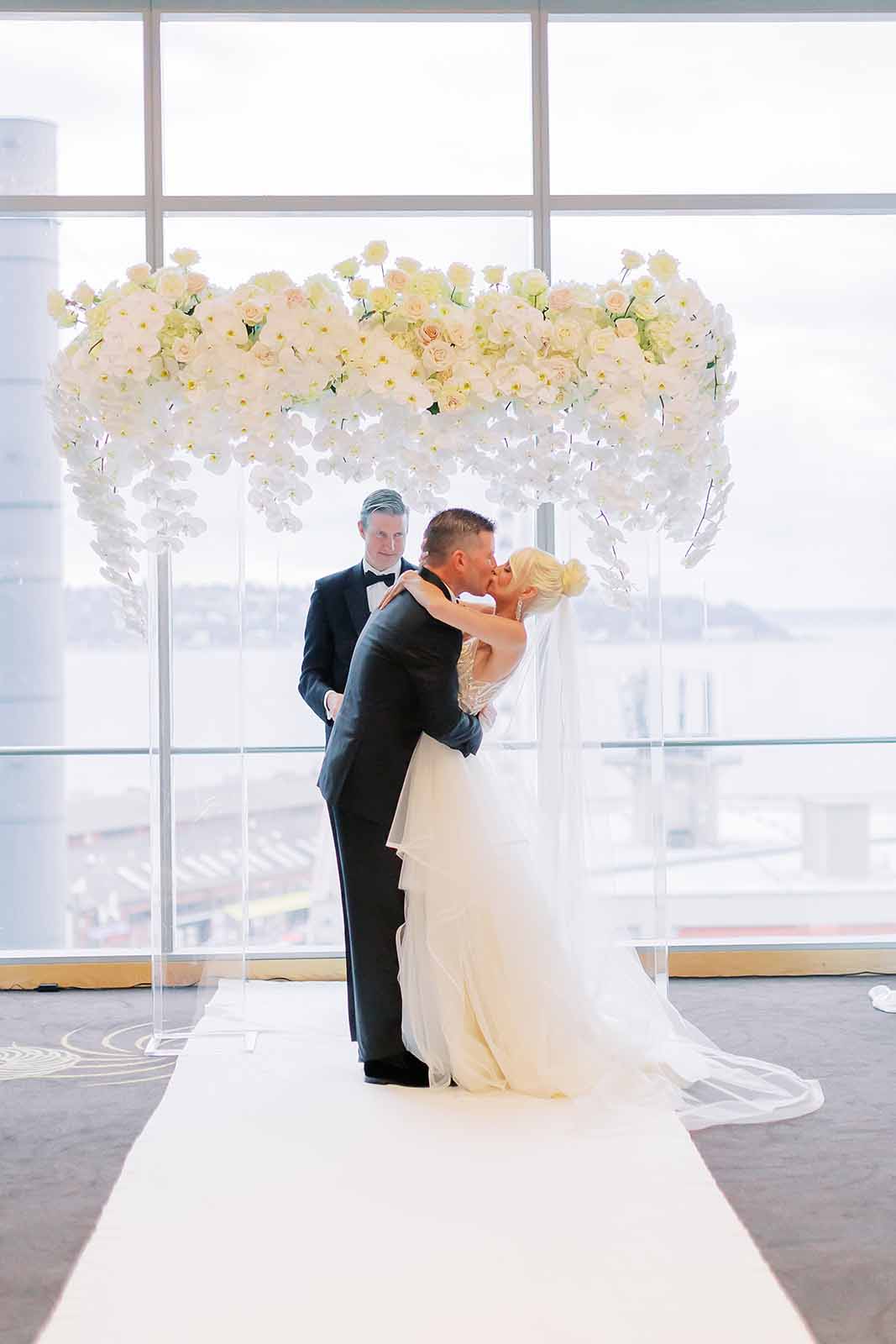 Bride and Groom kissing in front of a white floral arch covered in orchids during their formal ballroom wedding ceremony