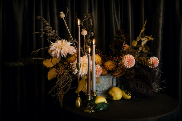 Moody centerpiece with orange dahlias, fall textures, stone vase, quince fruit, taper candles, on brown linen