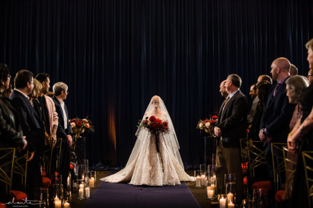 Bride with a red bouquet and full length veil makes a dramatic entrance to her winter ceremony lit by lots of candles lining the aisle | Flora Nova Design Seattle