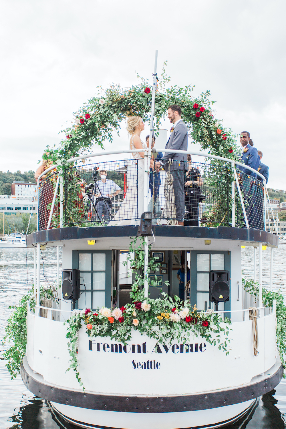 Bride and groom holding hand at fall wedding on Seattle's Lake Union with greenery arch and socially distanced guests - a safe pandemic wedding in Seattle