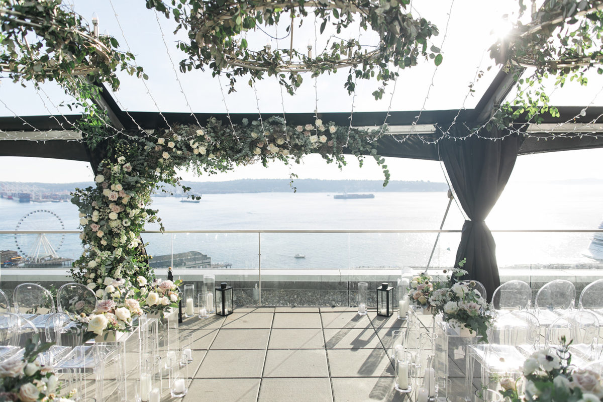 Elegant Seattle rooftop wedding with view of the Puget Sound: large greenery arch with white flowers, lots of candles and lanterns, and white flowers on clear lucite pedestals