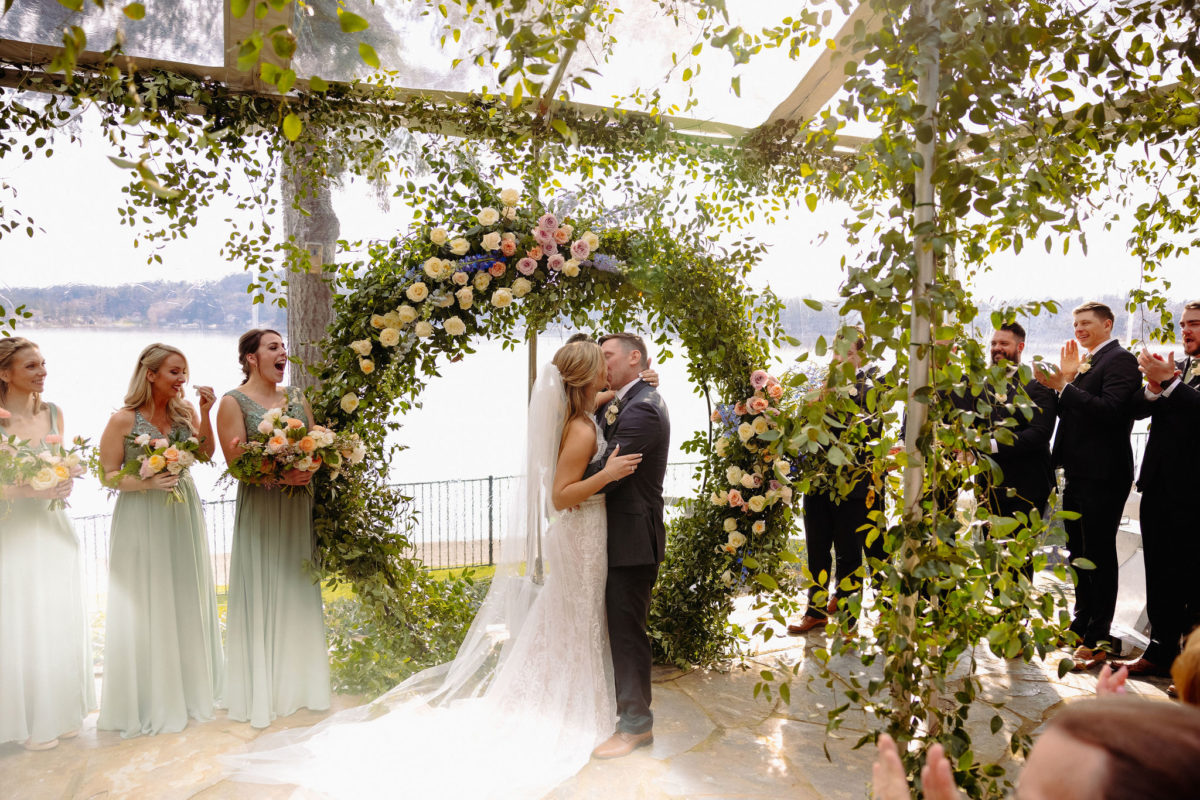 First kiss as husband and wife on their wedding day in a tent decorated with greenery and flowers, with the shores of Puget Sound in the background