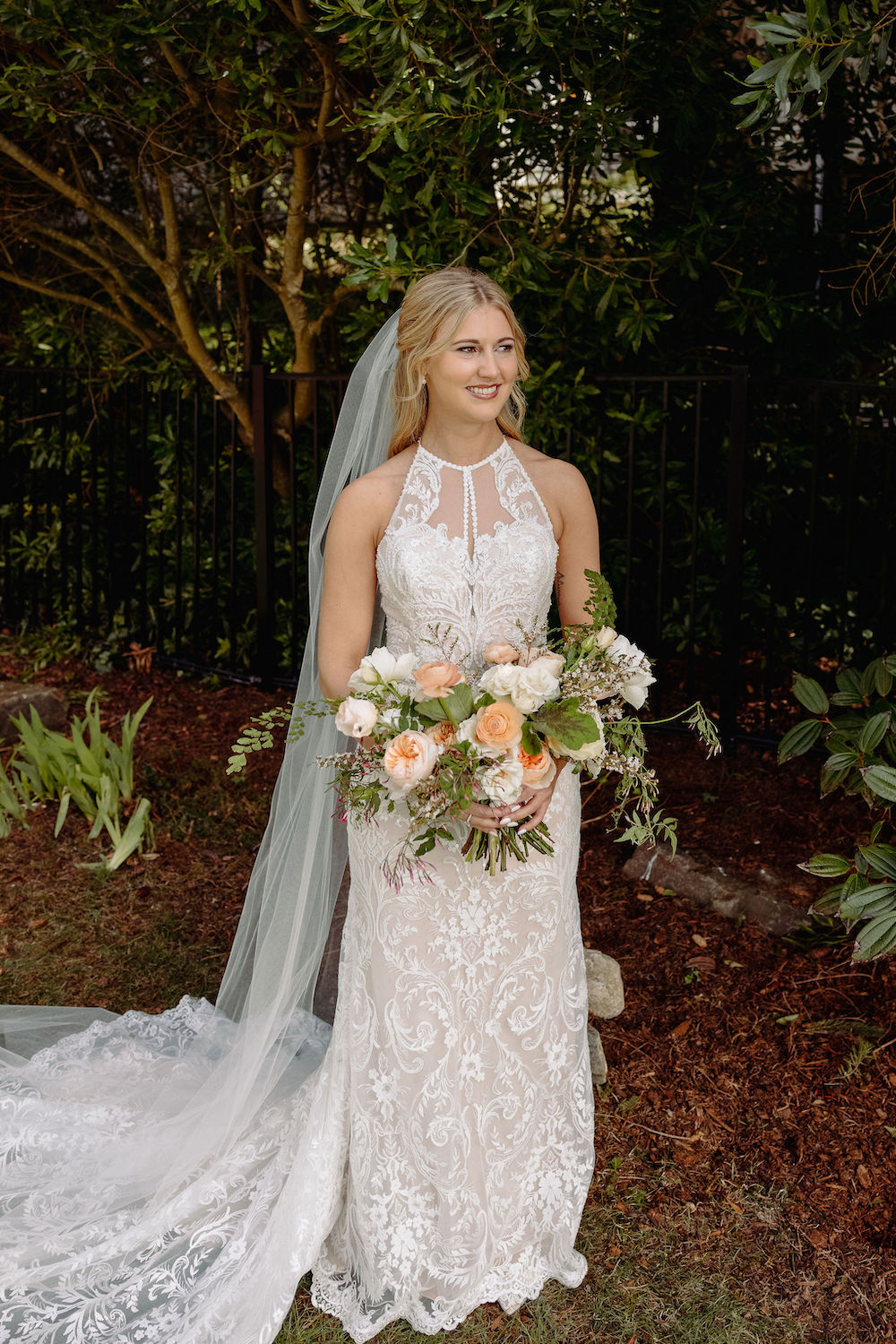 Bride in her long lace dress with a long veil holding her bouquet of peach garden roses, peach ranunculus, white spray roses, jasmine vine, cream roses, and geranium foliage