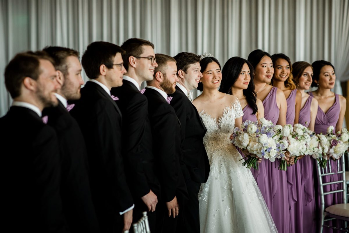 Wedding party with groomsmen in black tuxedos and bridesmaids in purple dresses holding white and purple spring bouquets