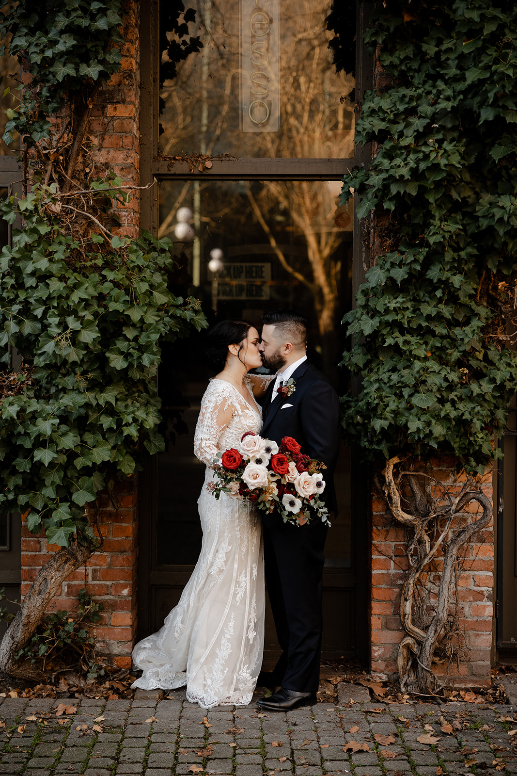 Bride and groom kiss standing amongst overgrown trees between brick walls.