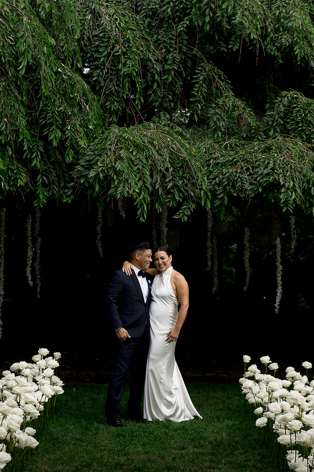 Bride and groom under a lush ceremony tree with white roses on either side of the aisle.
