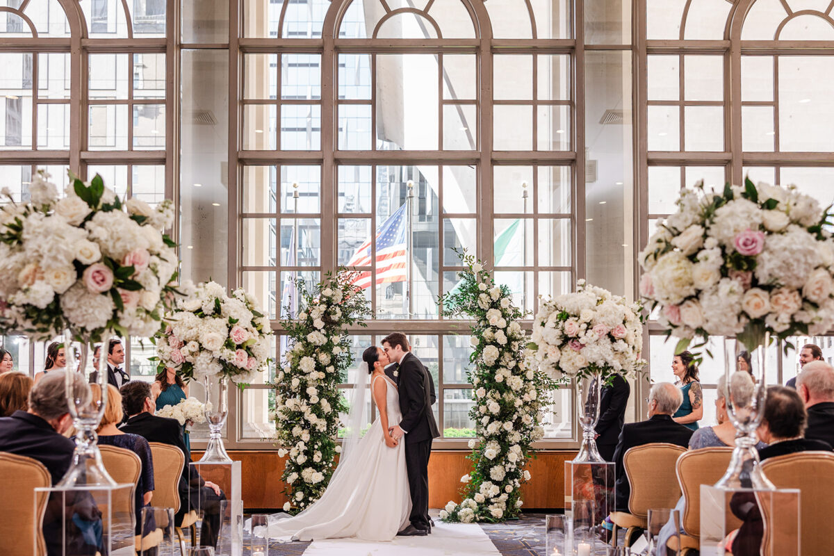 The bride and groom kiss at the end of the ceremony in for too the charming white and blush arch.
