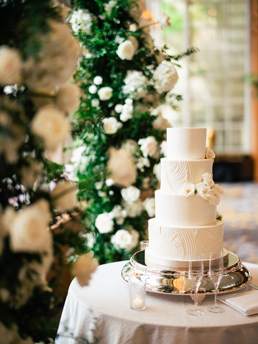 A white wedding cake and champagne glasses are placed by the lush green and floral arch