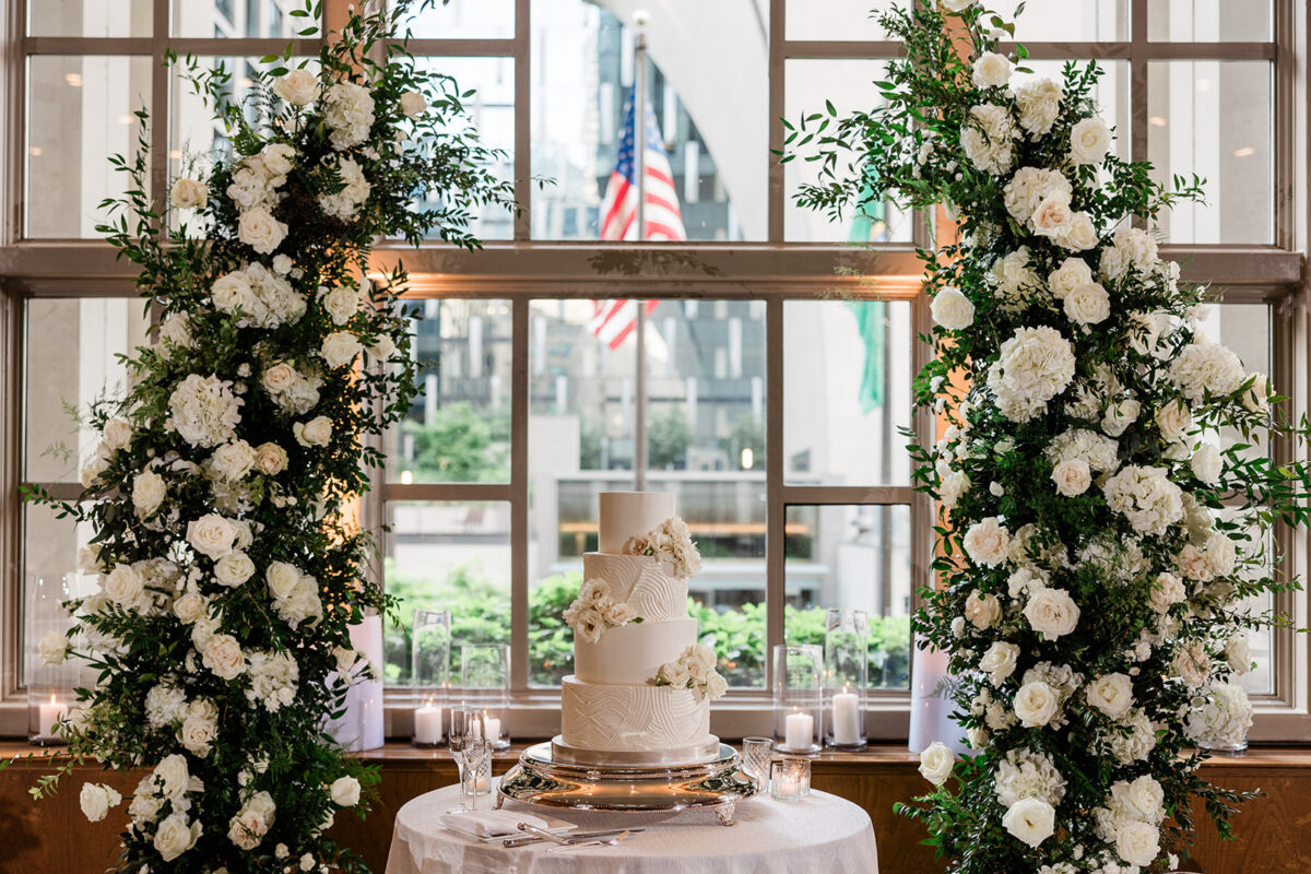 The cake table is places under the lush arch designed with white and blush florals and greenery.