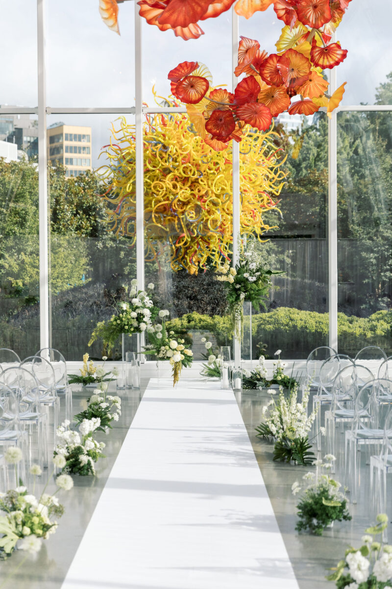 Chihuliy Glass House Aisle lined with ghost chairs and whimsical moss garden arrangements.
