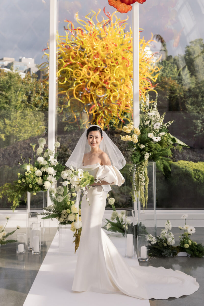 The bride stands in front of the ceremony space with her bouquet surrounded by whimsical moss garden arrangements.