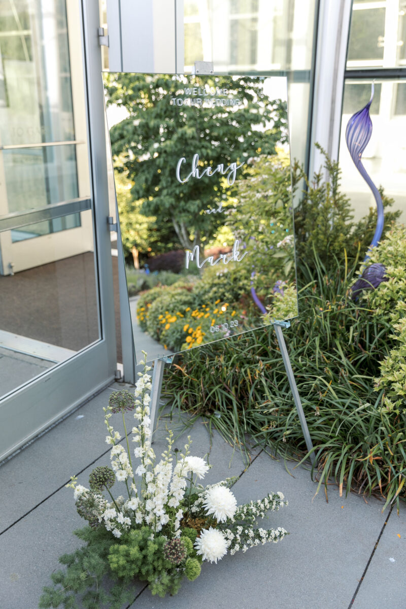 A mirrored welcome sign reflects the greenery at Chihuly Gardens and a floral arrangement sits at the foot of the sign.