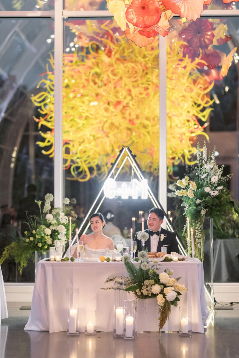 The bride and groom smile and sit at the sweetheart table surrounded by whimsical moss garden arrangements, candles, and neon lights.
