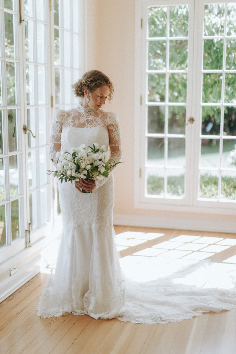 Bride holds her lush white bridal bouquet as she stands in the summer sun in her lace wedding dress.