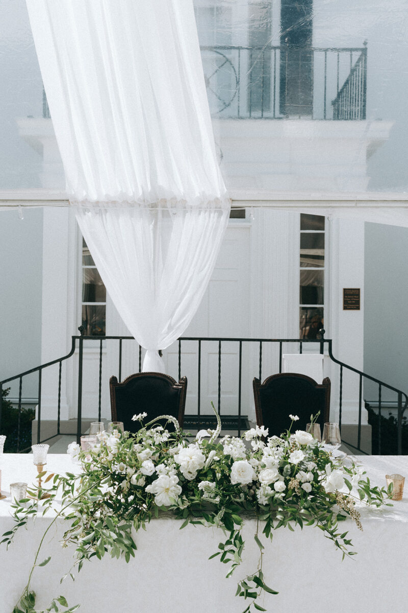 The lovely sweetheart table is decorated with a timeless white floral arrangement with growing greenery and white roses.