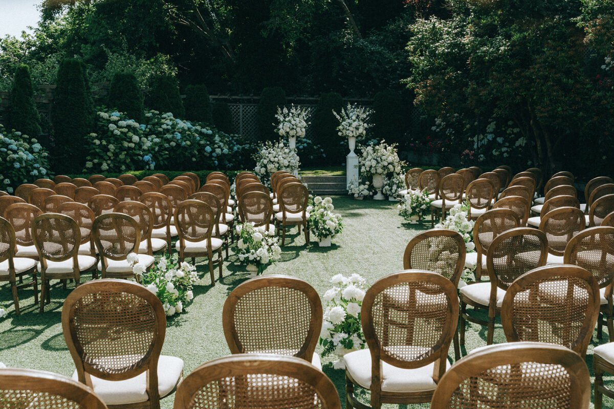 The soft curved aisle is lined with lush white arrangements and rows of brown wicker chairs for guests.