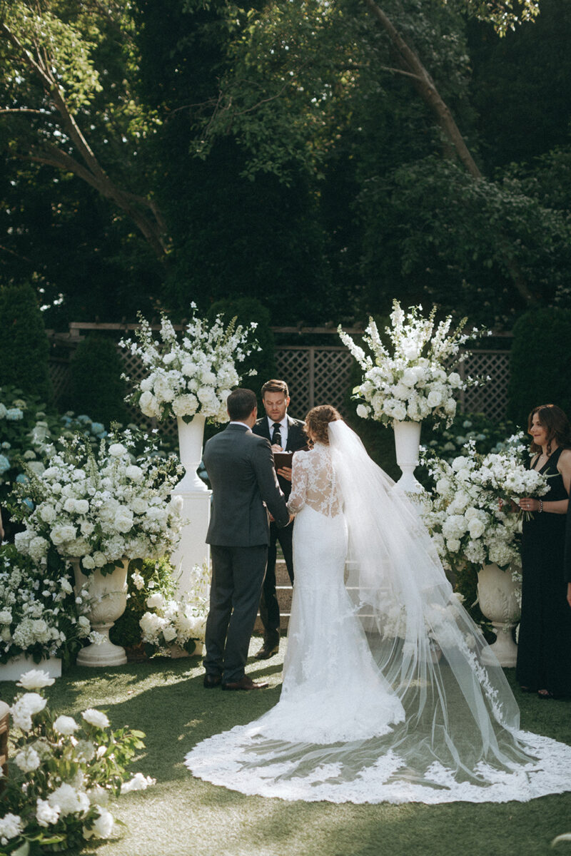 Bride and groom hold hands during the ceremony, surrounded by lush white floral arrangements.