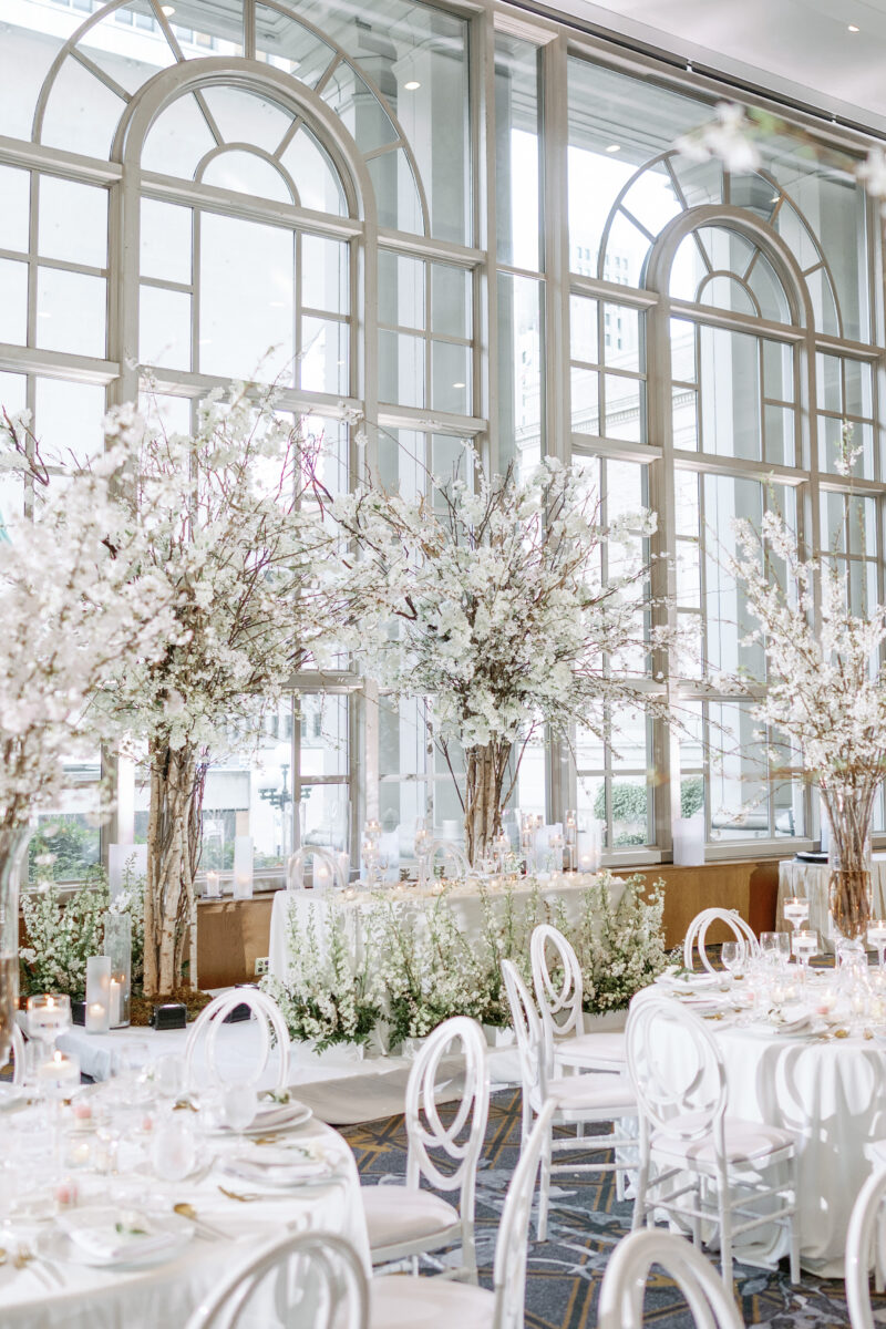 The sweetheart table is decorated with two cherry blossom trees and a meadow of lush floral arrangements backlit by the floor to ceiling windows in the Garden room.