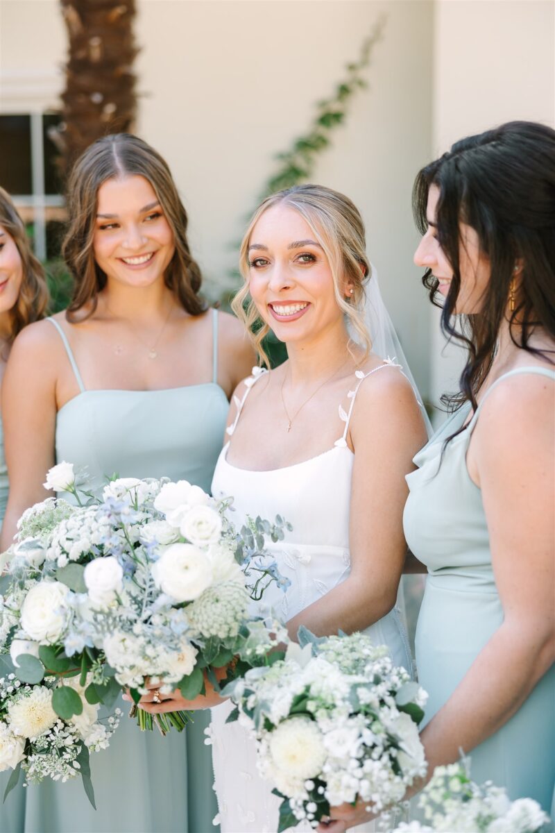 Bride smiles as she is surrounded by her bridesmaids holding her lush white and green bouquet with pops of blue.