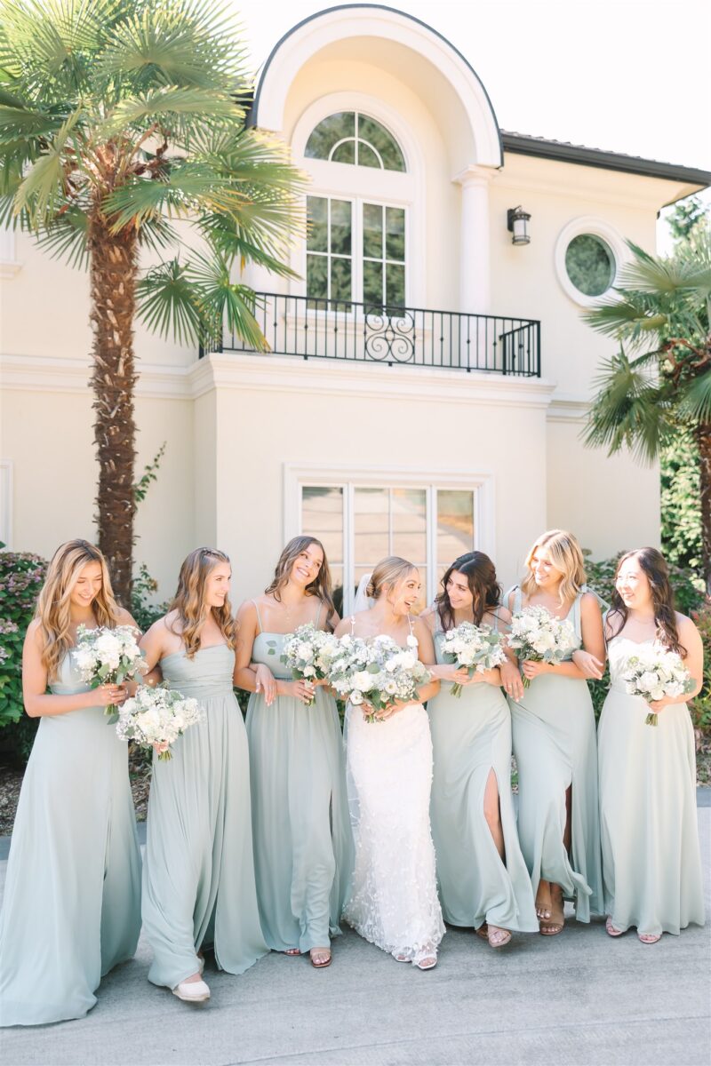 Bride and bridesmaids walk hand and hand with their lush bouquets in front of the private residence.