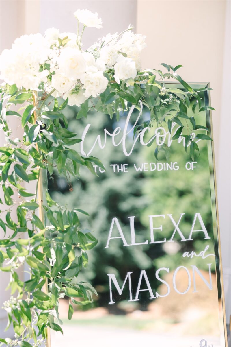 A mirror welcome sign is decorated with greenery and a lush white arrangement.