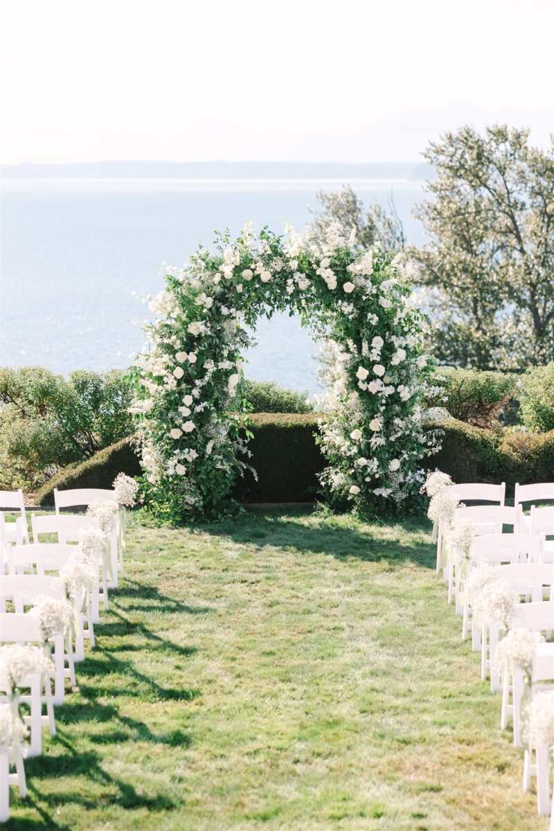 Aisle chairs with baby's breath ties and a lush white and green arch decorate the ceremony space.