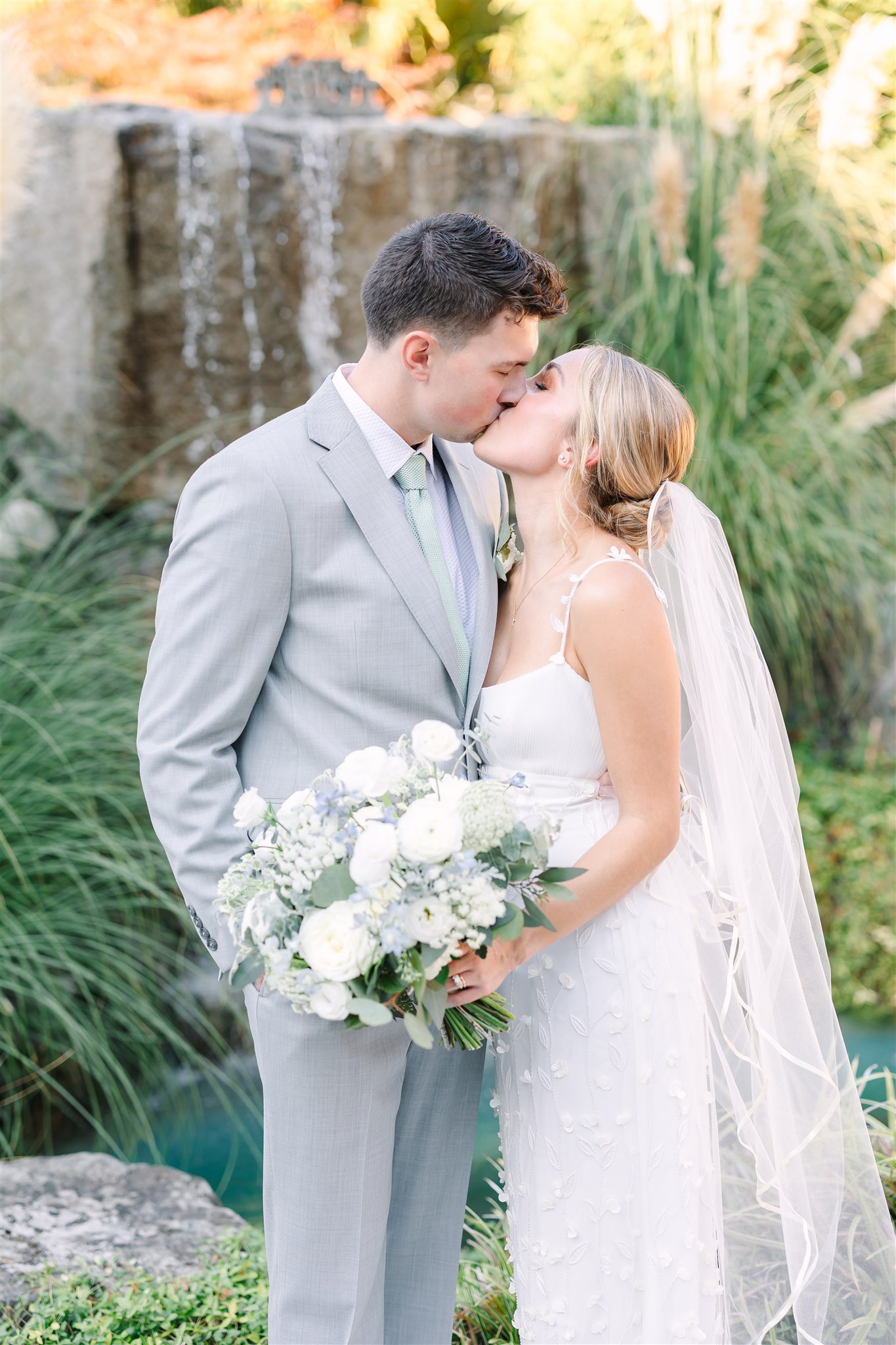 Bride and groom kiss in front of the waterfall at the glamorous wedding at a private residence.
