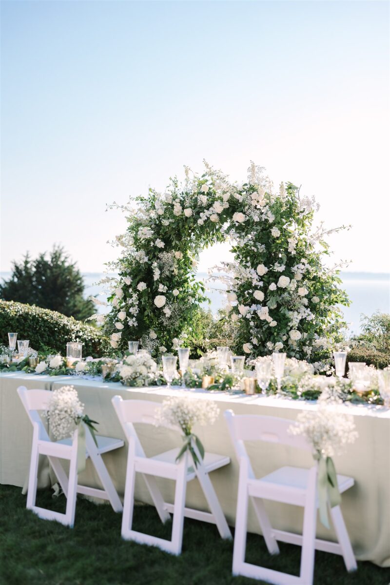 The head table is filled with white floral arrangements and votive candles with the arch as a backdrop to the bride and grooms seats.