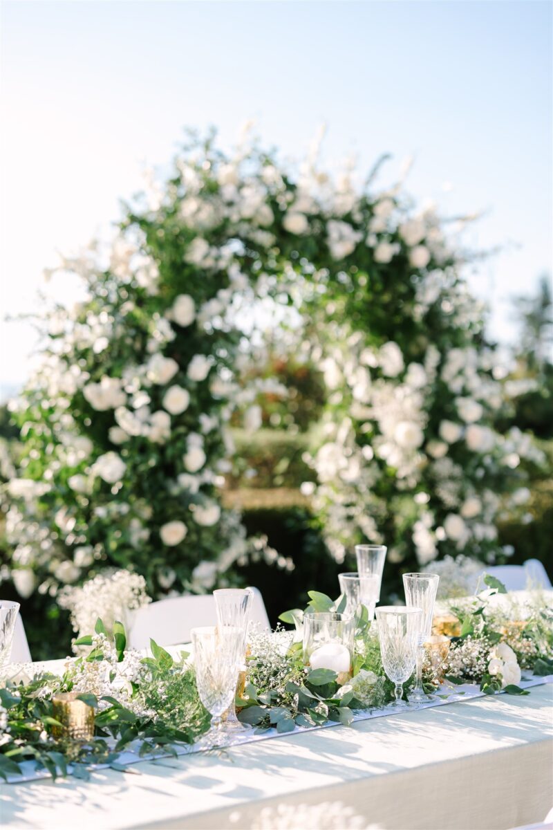 The head table at the glamorous wedding at a private residence is filled with greenery and florals.