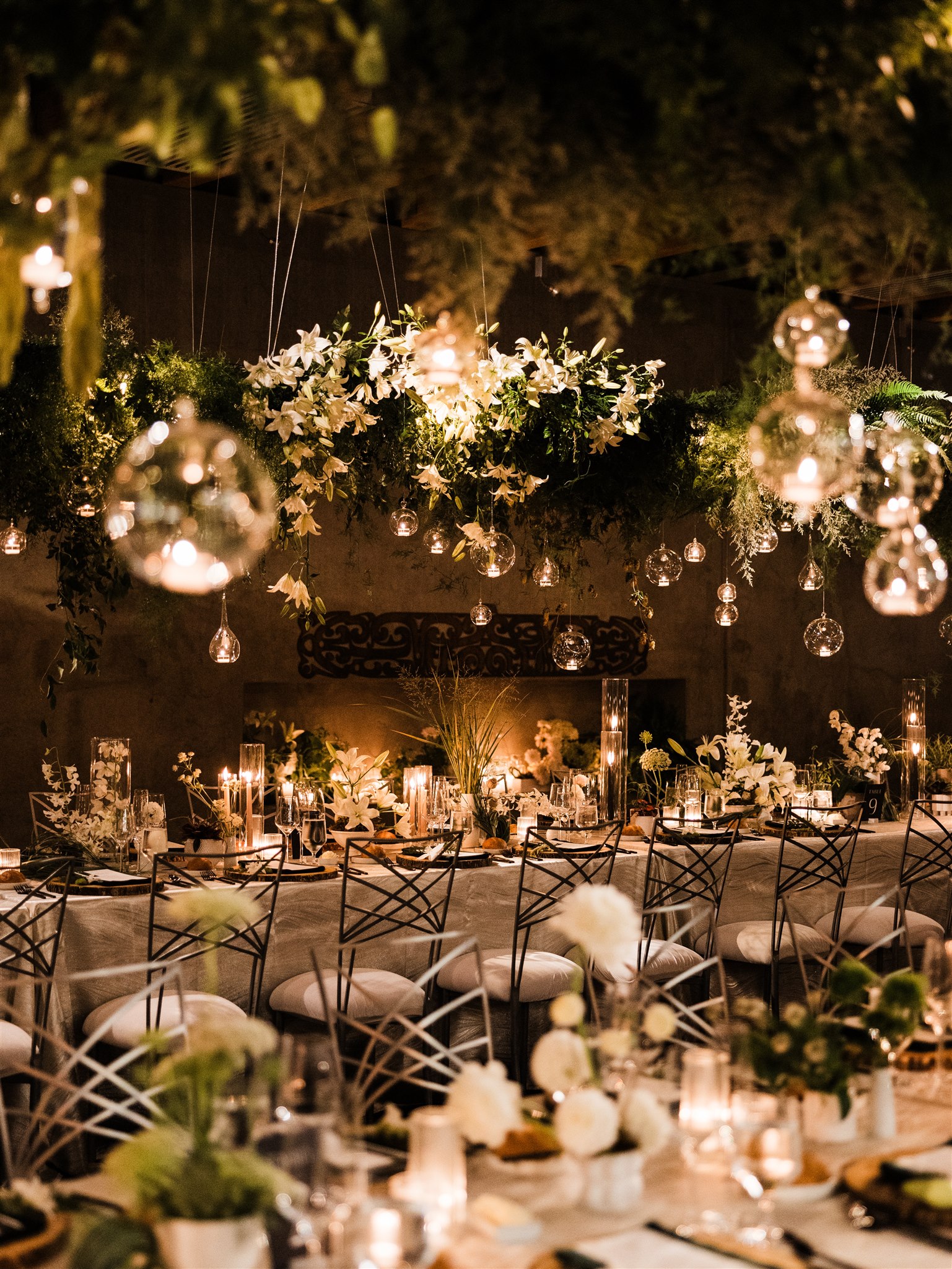 Long dining tables at wedding reception with hanging ceiling greenery accented by hanging round candles and white lilies suspended from the greenery.