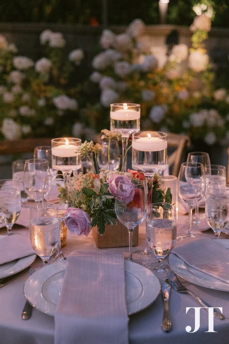 Evening centerpiece detail with floating candles in stemware, purple ranunculus, and mixed seasonal blooms creating romantic atmosphere at Lake Washington venue