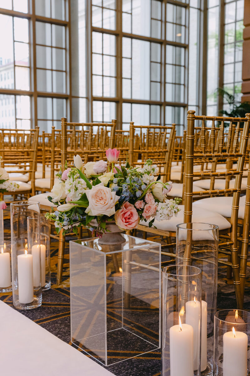 Sophisticated wedding ceremony setup with transparent acrylic pedestals displaying romantic floral arrangements of blush roses and white blooms, complemented by flickering pillar candles in glass hurricanes
