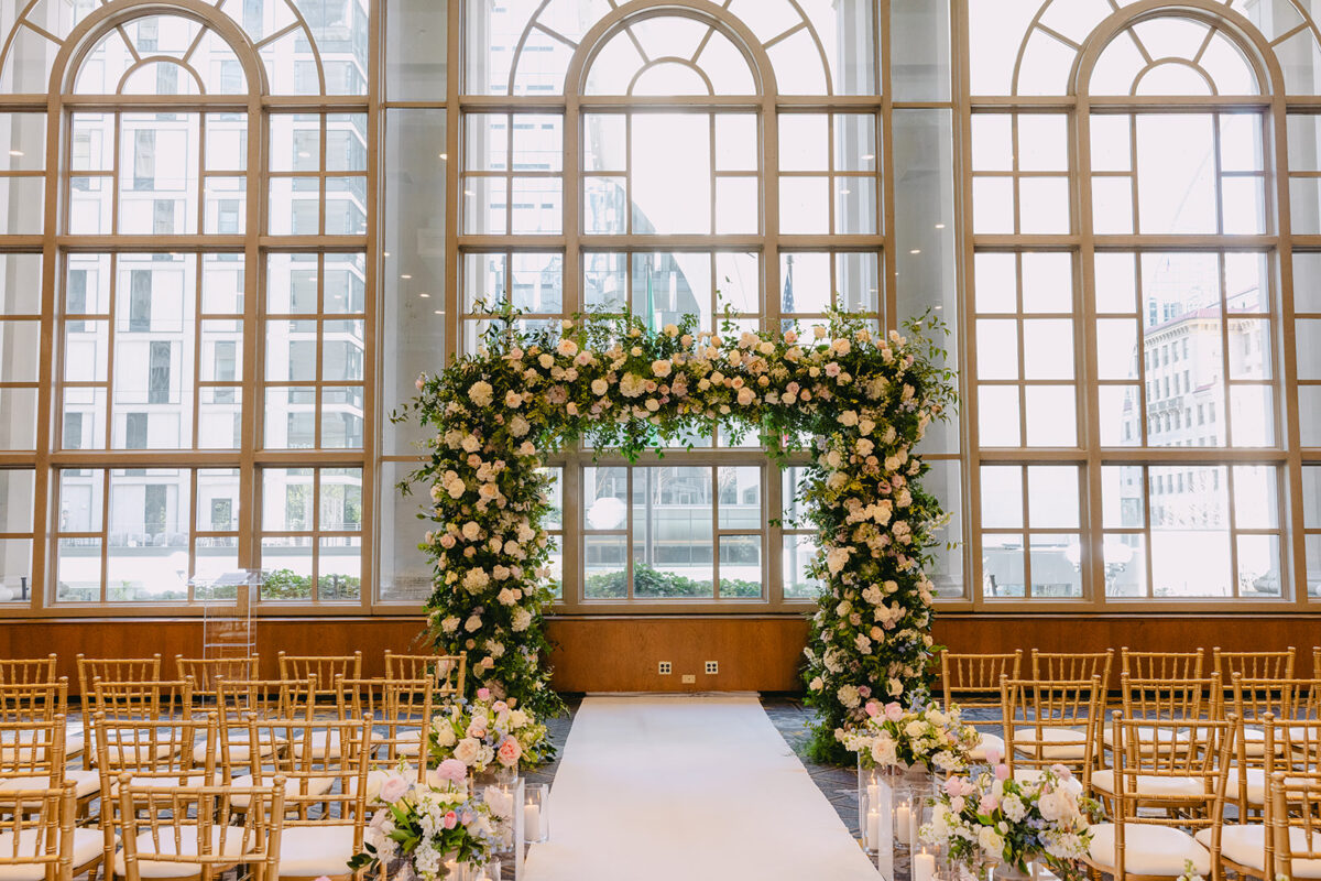 Stunning floral arch creates the perfect backdrop for this timeless wedding in Seattle, featuring cascading white and blush roses against floor-to-ceiling arched windows with city views