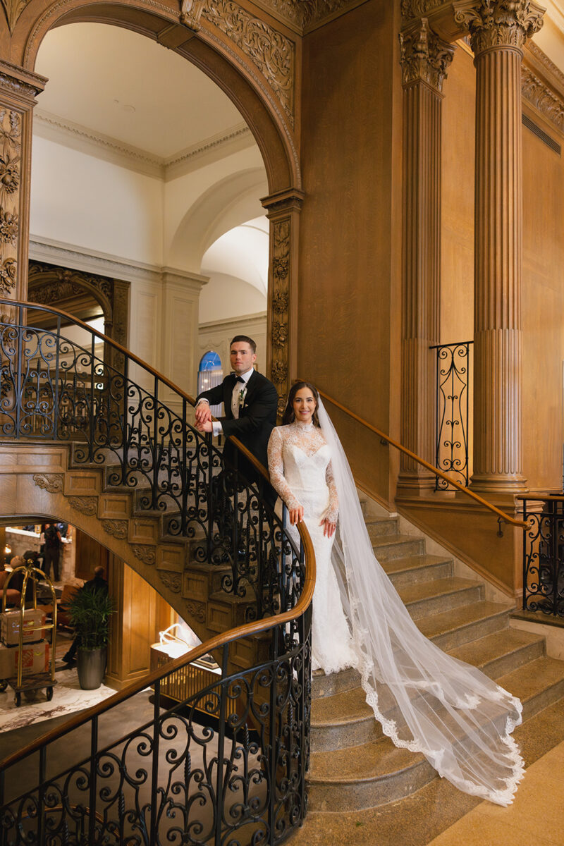 Bride and groom pose on grand marble staircase with ornate ironwork railings, bride wearing long-sleeved lace gown with cathedral veil