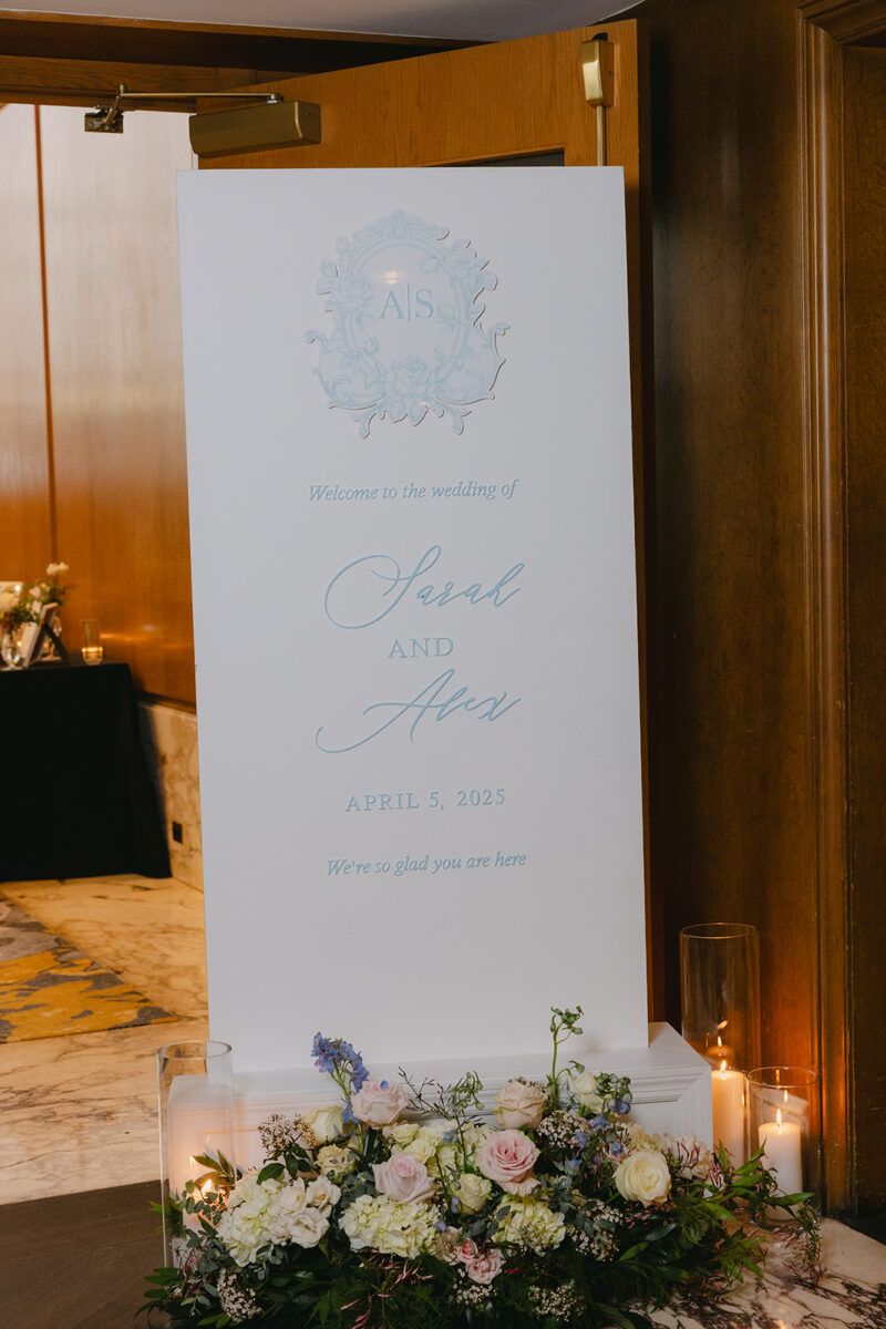 Personalized welcome sign featuring custom monogram and soft blue lettering, surrounded by delicate floral arrangements