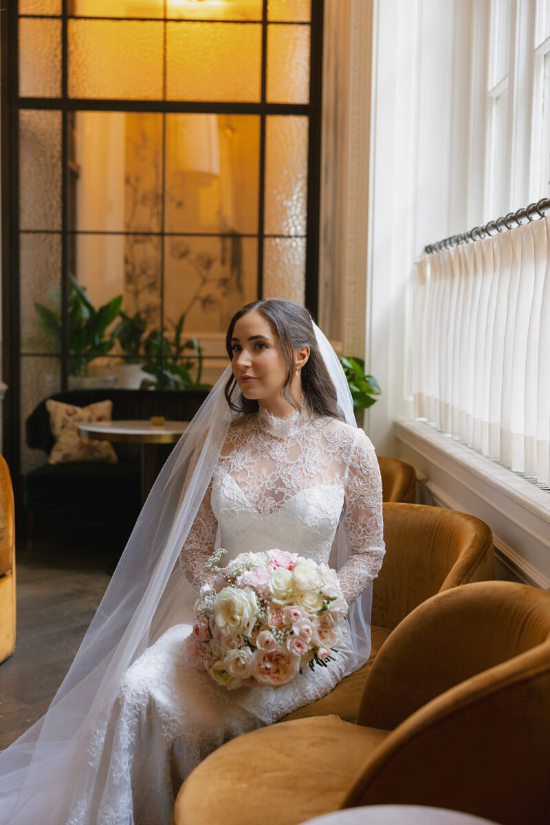 Radiant bride in long-sleeved lace wedding gown with fingertip veil, holding a classic bouquet of white and blush roses while seated by large arched windows