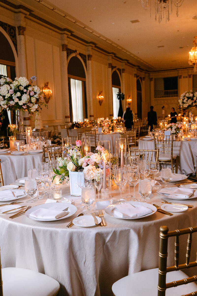 Classic reception table detail showing low garden-style arrangements of blush roses and white blooms, surrounded by crystal glassware and gold accents in the candlelit ballroom