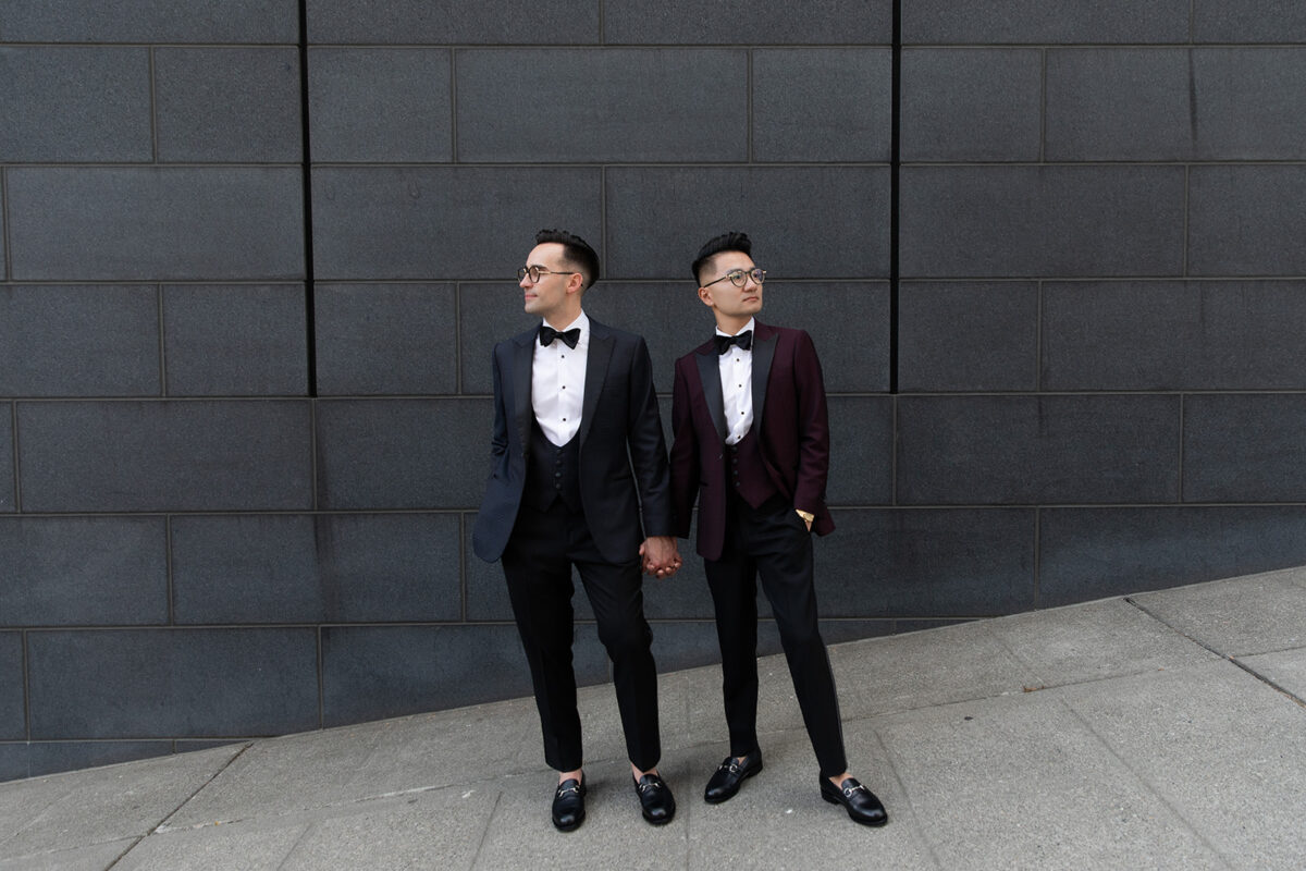 Two grooms in formal tuxedos standing hand-in-hand against modern gray wall before their Elegant Hotel Lotte Wedding ceremony