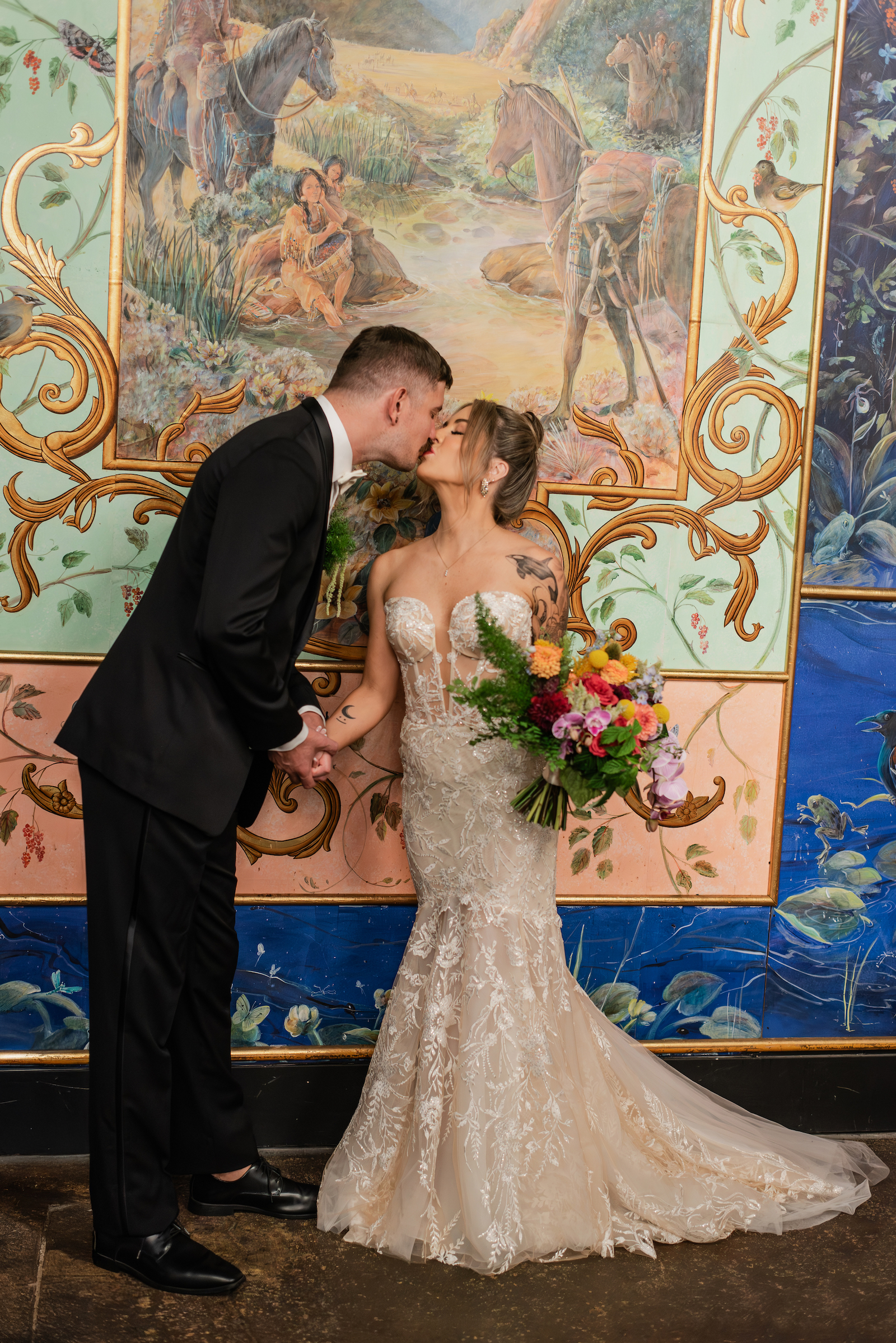 A colorful wedding at The Ruins by Flora Nova Design Seattle. Bride and groom kissing in front of eclectic art piece. Bride is holding a colorful bouquet with a selection of bright flowers and accents of purple orchids.