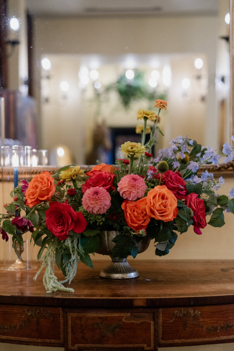 A decadent accent floral arrangement highlighted by blue taper candles. Flowers included in the compote arrangement were blu belladonna, dahlias, orange roses, pink roses and pops of yellow zinnias.